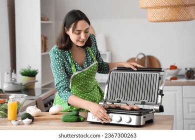 Beautiful young woman cooking delicious sausages on modern electric grill in kitchen - Shutterstock ID 2367300263