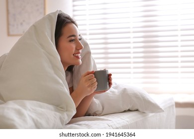 Beautiful young woman covered with warm blanket enjoying hot drink on bed at home. Space for text