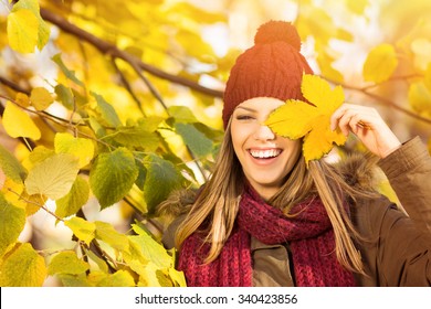 Beautiful Young Woman Covering Her Eye With Autumn Leaf Enjoying Autumn Standing Among Leaves In Treetop. Portrait Of Happy Teenage Girl In Red Beanie And Scarf In Park In Fall. Retouched, Vibrant.