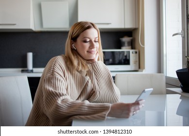 beautiful young woman in a cozy sweater sitting in the kitchen uses subscription services and media applications. A girl at home with a phone in her hands and wireless headphones in her ears.