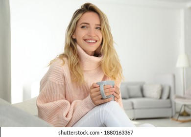 Beautiful young woman with cup of hot drink wearing warm pink sweater at home