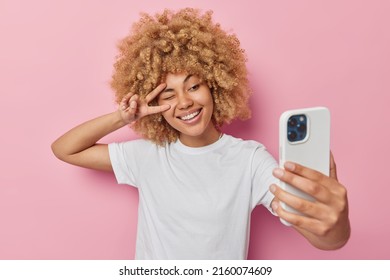 Beautiful young woman with curly blonde hair makes peace gesture over eye poses at smartphone camera dressed in casual white t shirt isolated over pink studio background talks on video call app - Powered by Shutterstock