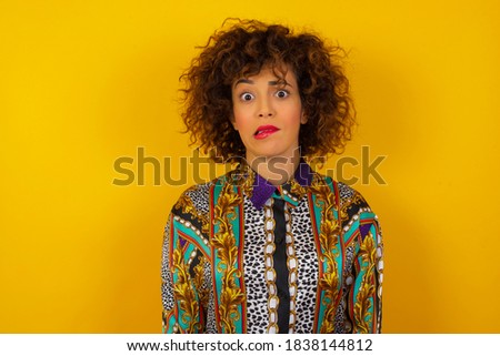 Beautiful young  woman with curly hair wearing colorful shirt over yellow background nervous and scared biting lips looking camera with impatient expression, pensive.