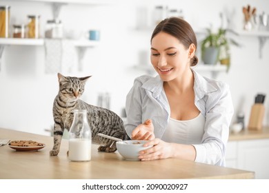 Beautiful young woman with cute cat eating cornflakes in kitchen