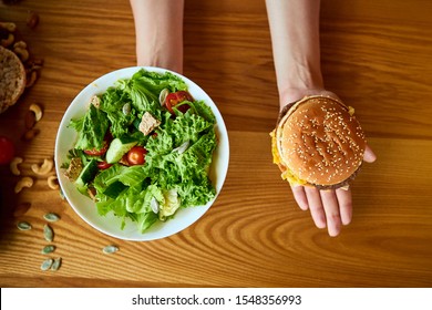 Beautiful Young Woman Decides Eating Hamburger Or Fresh Salad In Kitchen. Cheap Junk Food Vs Healthy Diet