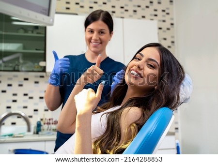 Beautiful young woman with dental braces sitting in dentist chair and her and her doctor showing thumbs up 