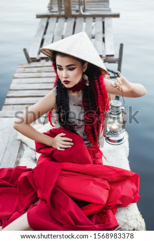 beautiful young woman in designer clothes and hat in asian style, with black hair and red braids, is sitting on the wooden bridge on the river with a vintage lantern