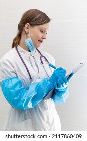 Beautiful Young Woman Doctor In A Medical Gown, Mask And Blue Gloves With A Stethoscope Writes Down A Medical History In A Hospital. Selective Focus. Portrait. Close-up