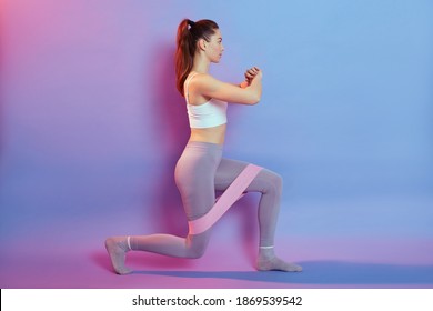 Beautiful young woman doing lunge exercise with sport fitness rubber bands isolated over pink and blue background. Fit girl living an active lifestyle, girl with perfect body wearing bra and leggins.