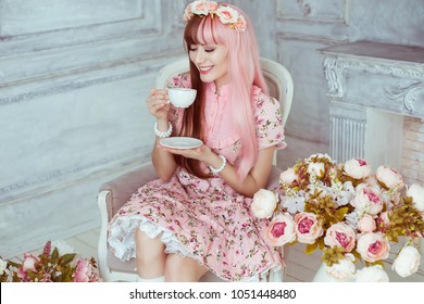 Beautiful young woman doll in a pink dress, in a bright room and flowers sitting in a chair with a cup, tea party, lolita. Japanese street fashion. Portrait.