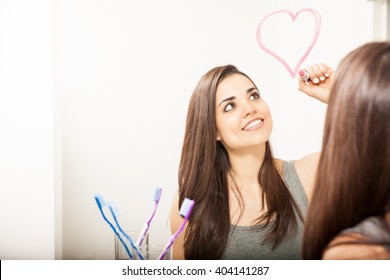 Beautiful young woman drawing heart in bathroom mirror using lipstick
