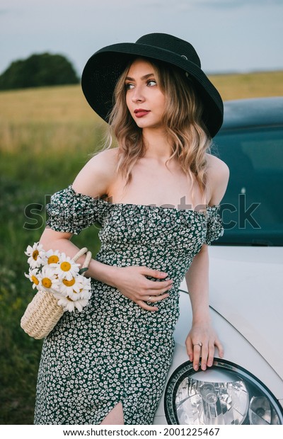 Beautiful young woman in dress and hat stands\
 \
near a white car.\
toning