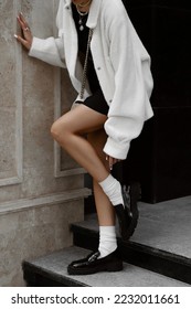 beautiful young woman dressed in black skirt and top, white furry jacket, patent leather loafers, socks, white bag, bun hairstyle, stylish fashion outfit, full length lifestyle model