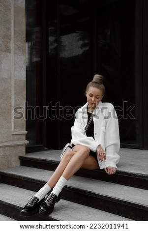 beautiful young woman dressed in black skirt and top, white furry jacket, patent leather loafers, socks, white bag, bun hairstyle, stylish fashion outfit, lifestyle model sitting