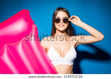 Beautiful young woman dressed in white swimsuit and glasses posing with pink inflatable mattress isolated over blue background