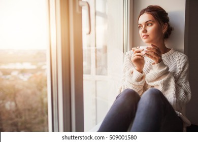 Schöne junge Frau trinkt Kaffee und sieht durch das Fenster, während sie zu Hause auf Fensterbrett sitzt