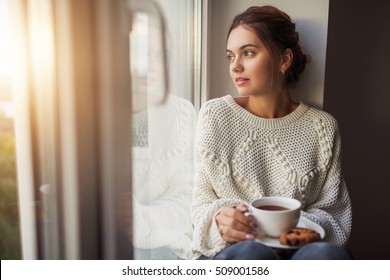 Schöne junge Frau trinkt Kaffee und sieht durch das Fenster, während sie zu Hause auf Fensterbrett sitzt