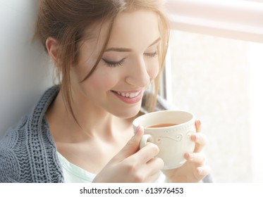 Beautiful young woman drinking tea near window at home