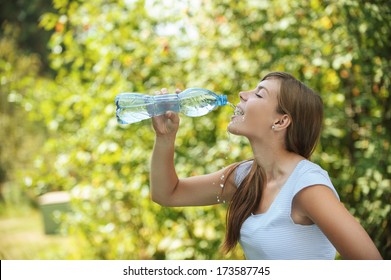 Beautiful young woman drinks water from bottle, against green of summer park.