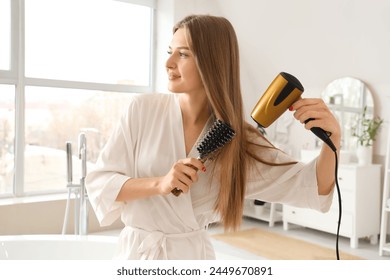 Beautiful young woman drying her hair with round brush in bathroom