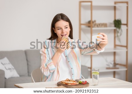 Beautiful young woman eating tasty quesadilla and taking selfie at home