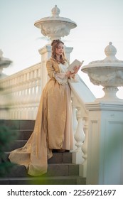 A beautiful young woman in an eighteenth century historical gold dress stands on the stairs of a mansion with a book in her hands. the princess is reading in the palace. fabulous image - Shutterstock ID 2256921697