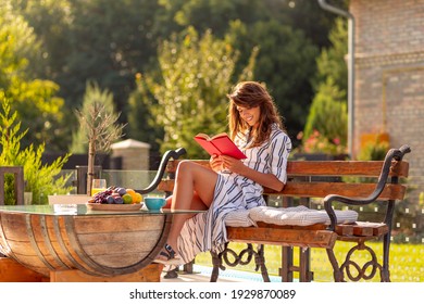 Beautiful Young Woman Enjoying Sunny Summer Morning Outdoors, Drinking Coffee And Reading A Book  On The Backyard Terrace