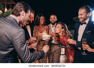 Beautiful young woman in evening gown blowing candles on cake while celebrating birthday with friends - Powered by Shutterstock