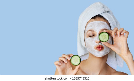 Beautiful Young Woman With Facial Mask On Her Face Holding Slices Of Fresh Cucumber. Skin Care And Treatment, Spa, Natural Beauty And Cosmetology Concept, Over Blue Background With Copy Space