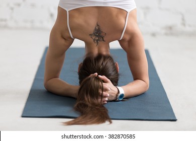 Beautiful young woman with flower tattoo on her back working out indoors, doing yoga exercise on blue mat, supported headstand posture, salamba sirsasana, rear view, close-up