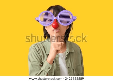 Beautiful young woman in funny disguise showing silent gesture on yellow background. April Fools Day celebration