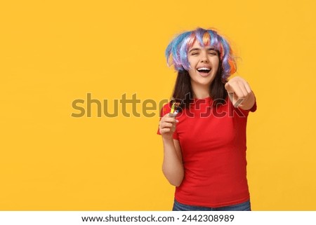 Beautiful young woman in funny disguise with party whistles pointing at viewer on yellow background. April fool's day celebration