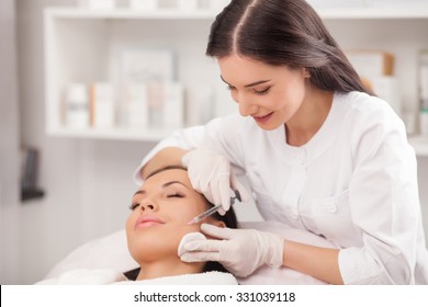 Beautiful young woman is getting botox injection in her cheek. She is lying with closed eyes. The doctor is holding syringe and smiling
