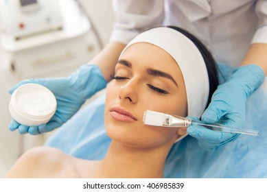 Beautiful Young Woman Is Getting Face Skin Treatment. Doctor In Medical Gloves Is Holding A Bottle Of Cream And A Brush