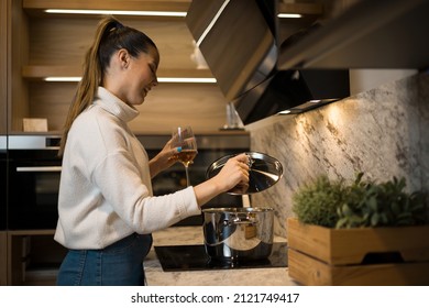 A Beautiful Young Woman With A Glass Of Wine In Her Hand Looking At The Soup She Is Cooking. Healthy Life Style.