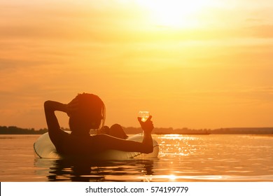Beautiful young woman with a glass of wine on inflatable ring in water during sunset