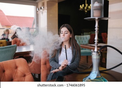 Beautiful young woman in grey jaket smoke hookah on terrace in restautant. Smoked