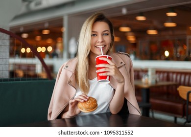 Beautiful young woman with a hamburger drinks a cola in a cafe - Shutterstock ID 1095311552
