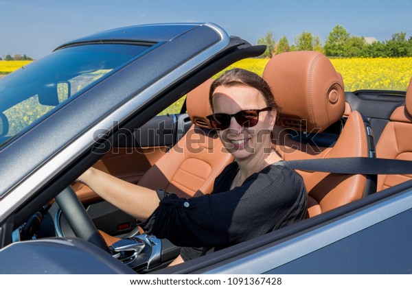 Beautiful young woman happy\
to drive her convertible car on a country road in summer. she looks\
at camera. 