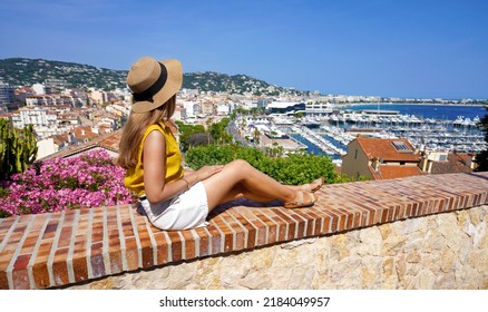 Beautiful Young Woman With Hat Sitting On Wall Looking At Cityscape Of Cannes, French Riviera