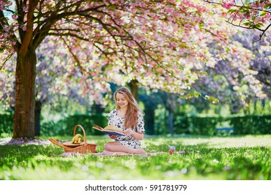 Beautiful young woman having picnic on sunny spring day in park during cherry blossom season, reading a book