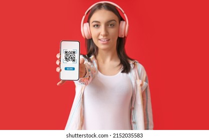 Beautiful young woman in headphones holding smartphone with QR code on screen against red background