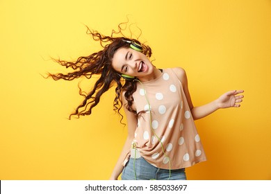 Beautiful young woman in headphones listening to music and dancing on yellow background