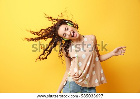 Beautiful young woman in headphones listening to music and dancing on yellow background