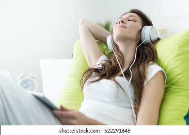 Beautiful Young Woman With Headphones Relaxing On The Bed, She Is Listening To Music Using A Tablet, Chill Out And Leisure Concept 