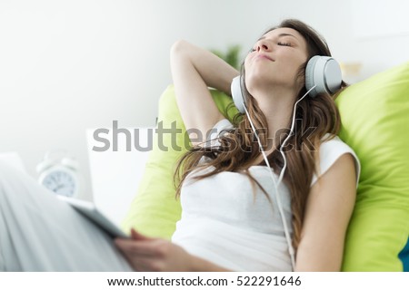 Beautiful young woman with headphones relaxing on the bed, she is listening to music using a tablet, chill out and leisure concept 