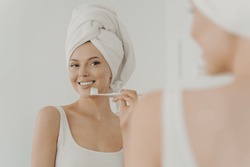 Beautiful Young Woman With Healthy Perfect Smile Brushing Teeth And Looking In Mirror, Attractive Young Female Wearing White Bath Towel On Head Standing In Bathroom At Home. Oral Hygiene Concept