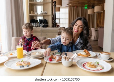 Beautiful Young Woman With Her Baby Sons. Light Breakfast Near Window In A Cafe. Croissants, Omelet, Coffee And Many Different Dishes On The Table In The Cafe. Happy Family