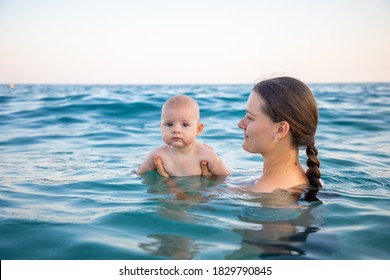 Beautiful Young Woman With Her Daughter Little Baby Girl Swimming In The Sea At Sunset Time. Woman With Daughter Relaxing In Front Of Sea