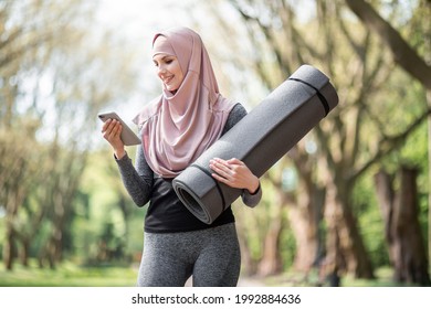 Beautiful Young Woman In Hijab Standing At Green Park With Yoga Mat And Using Modern Smartphone. Muslim Lady In Sport Clothes Resting During Workout In Fresh Air.